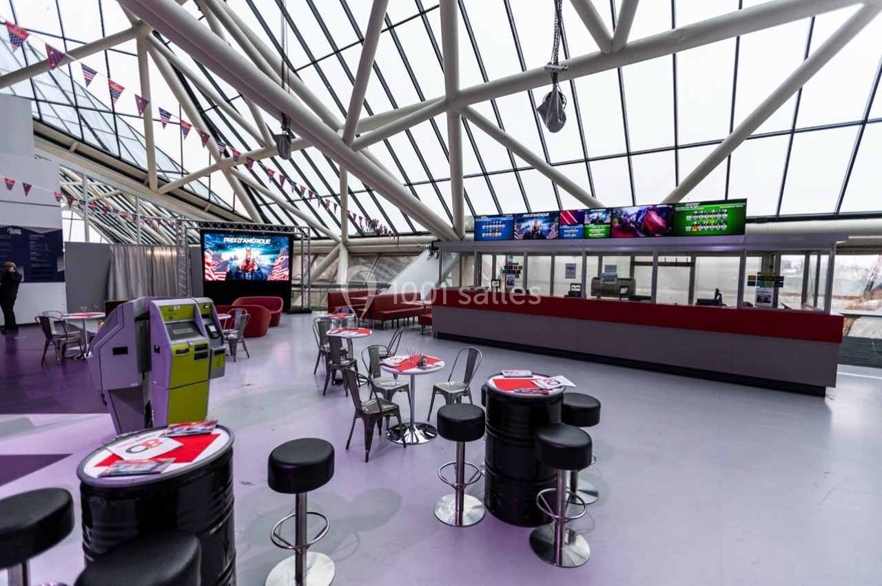Intérieur d'un espace moderne avec comptoirs, tables, chaises et écrans sous une verrière.