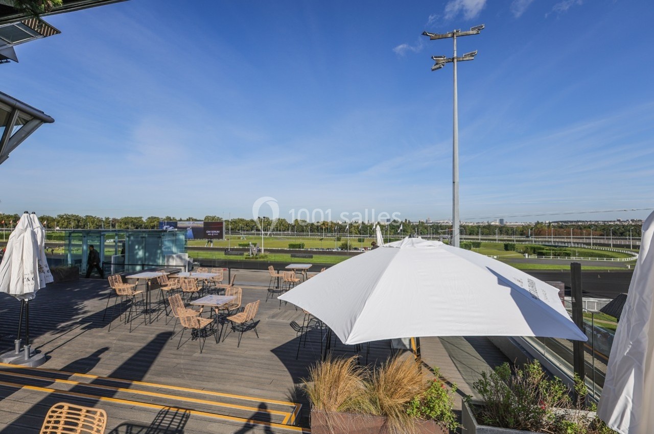 Terrasse en bois avec tables et parasols, vue sur un champ de course sous un ciel bleu dégagé.