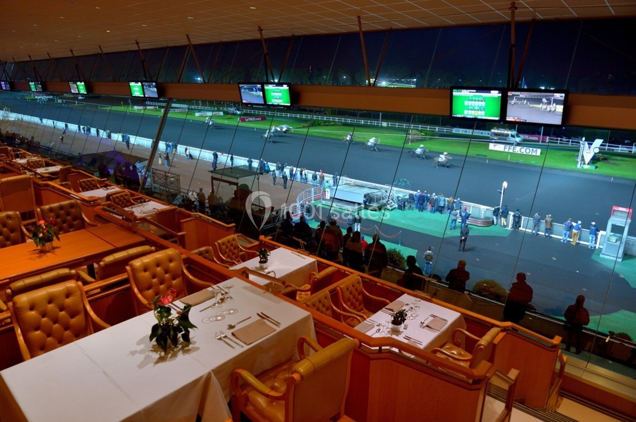 Vue depuis une salle de restaurant surplombant une piste de course hippique éclairée en soirée.
