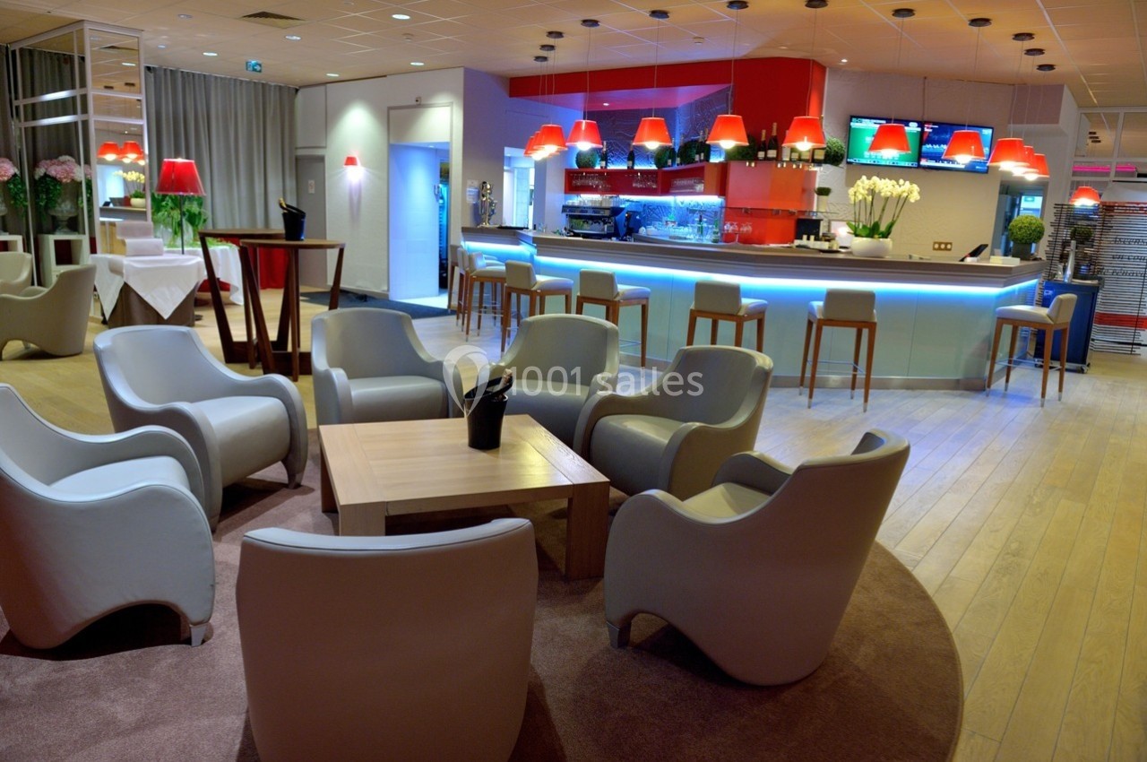 Salon moderne avec fauteuils gris, tables en bois clair et bar éclairé en arrière-plan, décoré de lampes rouges.