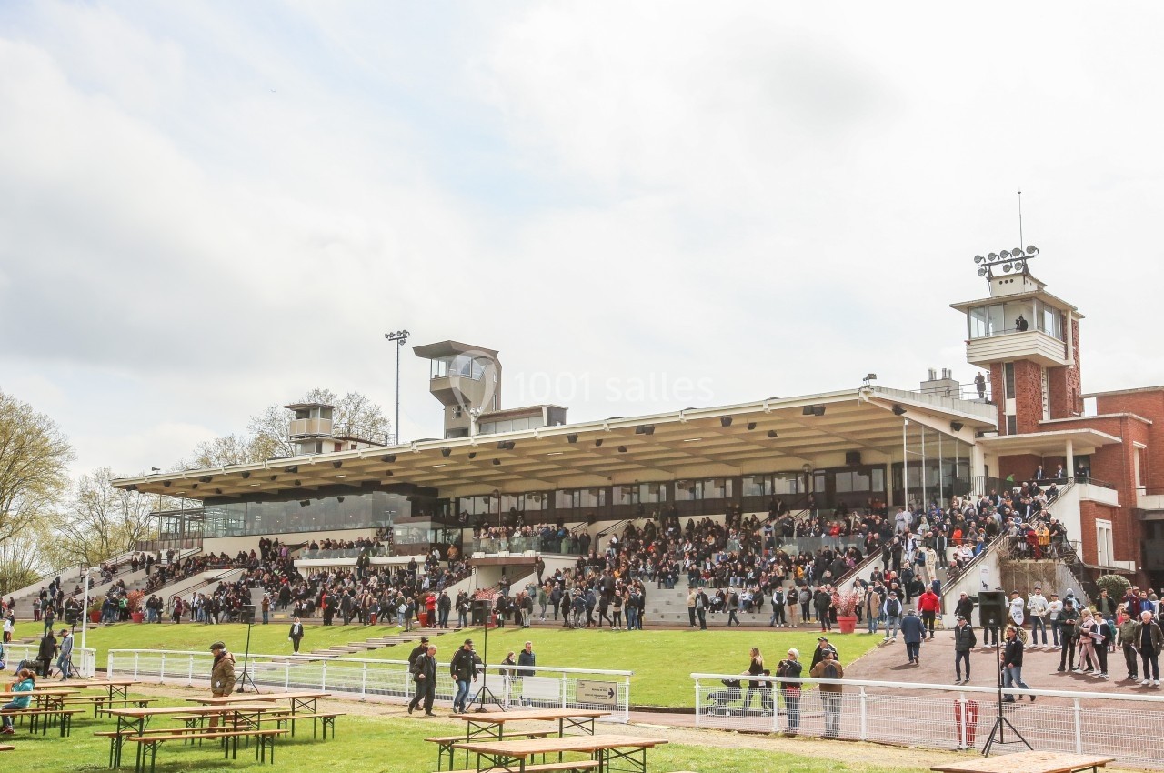 Tribune d'un hippodrome avec des spectateurs assis et debout, entourée d'espaces verts et de tables en plein air.