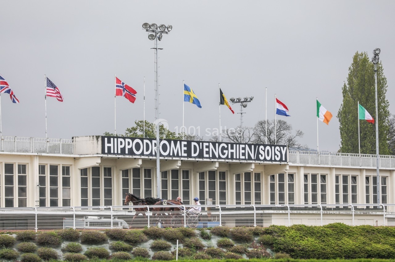 Façade de l'hippodrome d'Enghien-Soisy avec des drapeaux internationaux et un cheval attelé au premier plan.