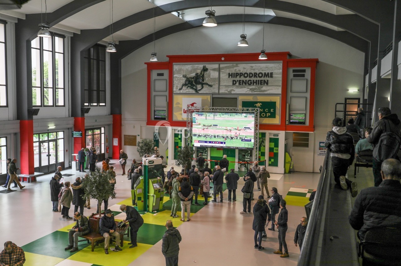 Vue intérieure d'un hall avec des écrans diffusant des courses hippiques, des spectateurs rassemblés au centre.