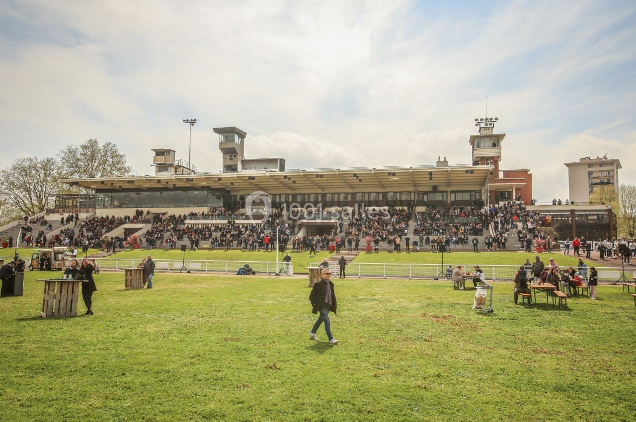 Tribune d'un hippodrome avec des spectateurs assis et debout, vue depuis une pelouse par temps clair.