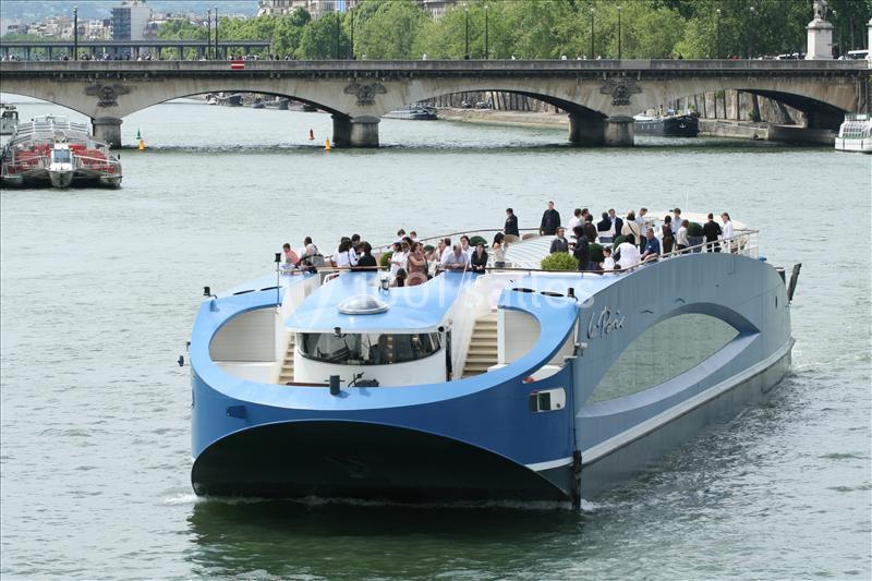 Bateau moderne transportant des passagers sur une rivière, avec un pont et des arbres en arrière-plan.