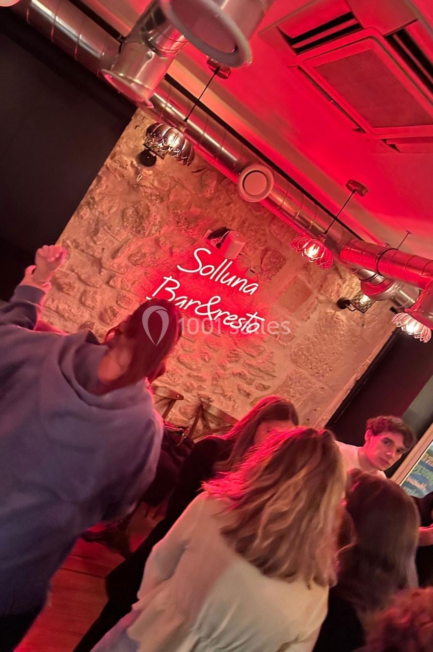 Des personnes discutent et dansent dans un bar à l'ambiance tamisée, avec un néon rouge indiquant ’Solluna Bar & Resto’.