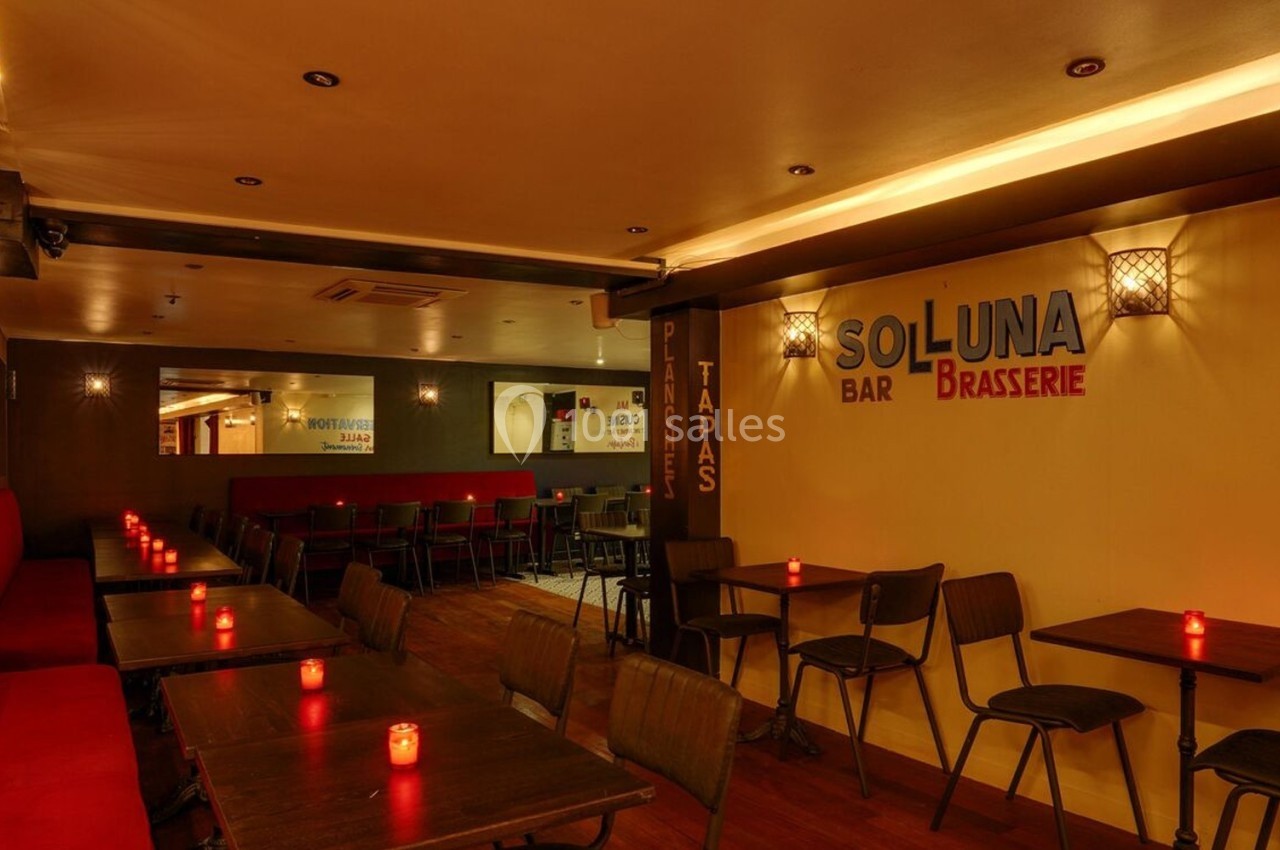 Salle de restaurant avec tables en bois, chaises noires, banquette rouge et bougies allumées pour une ambiance chaleureuse.
