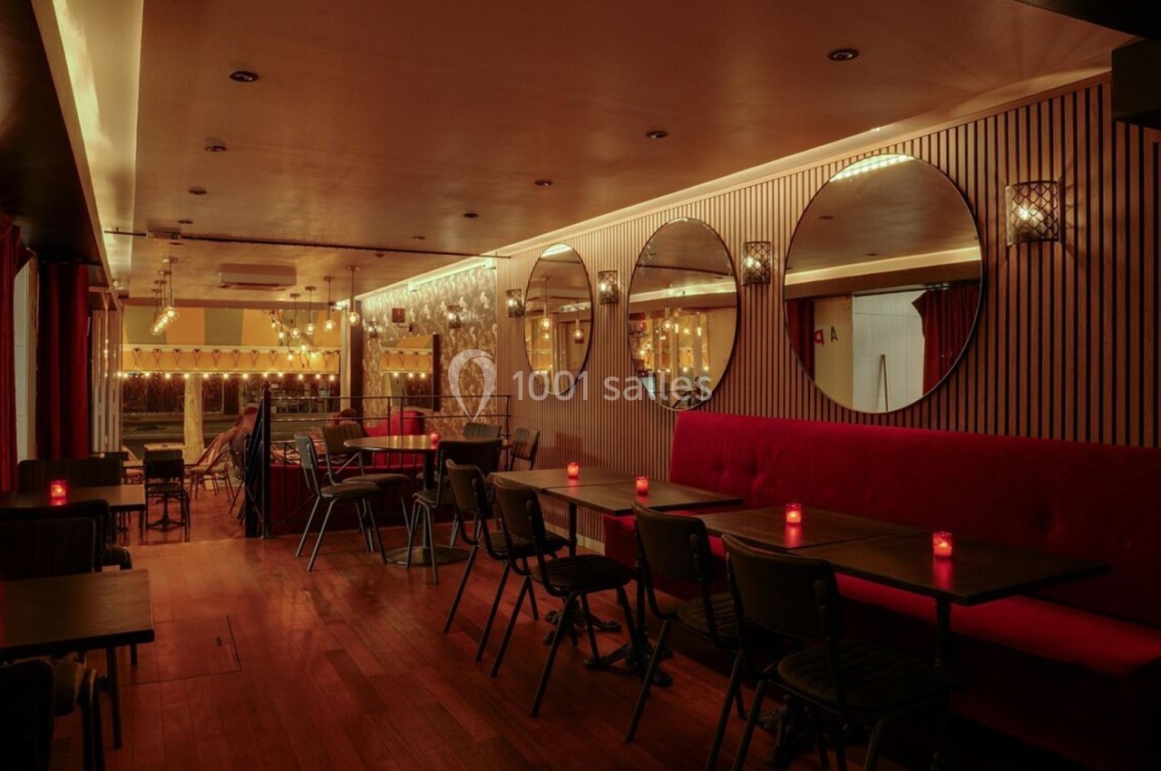 Salle de restaurant chaleureuse avec éclairage tamisé, banquettes rouges, miroirs ronds et petites tables noires.