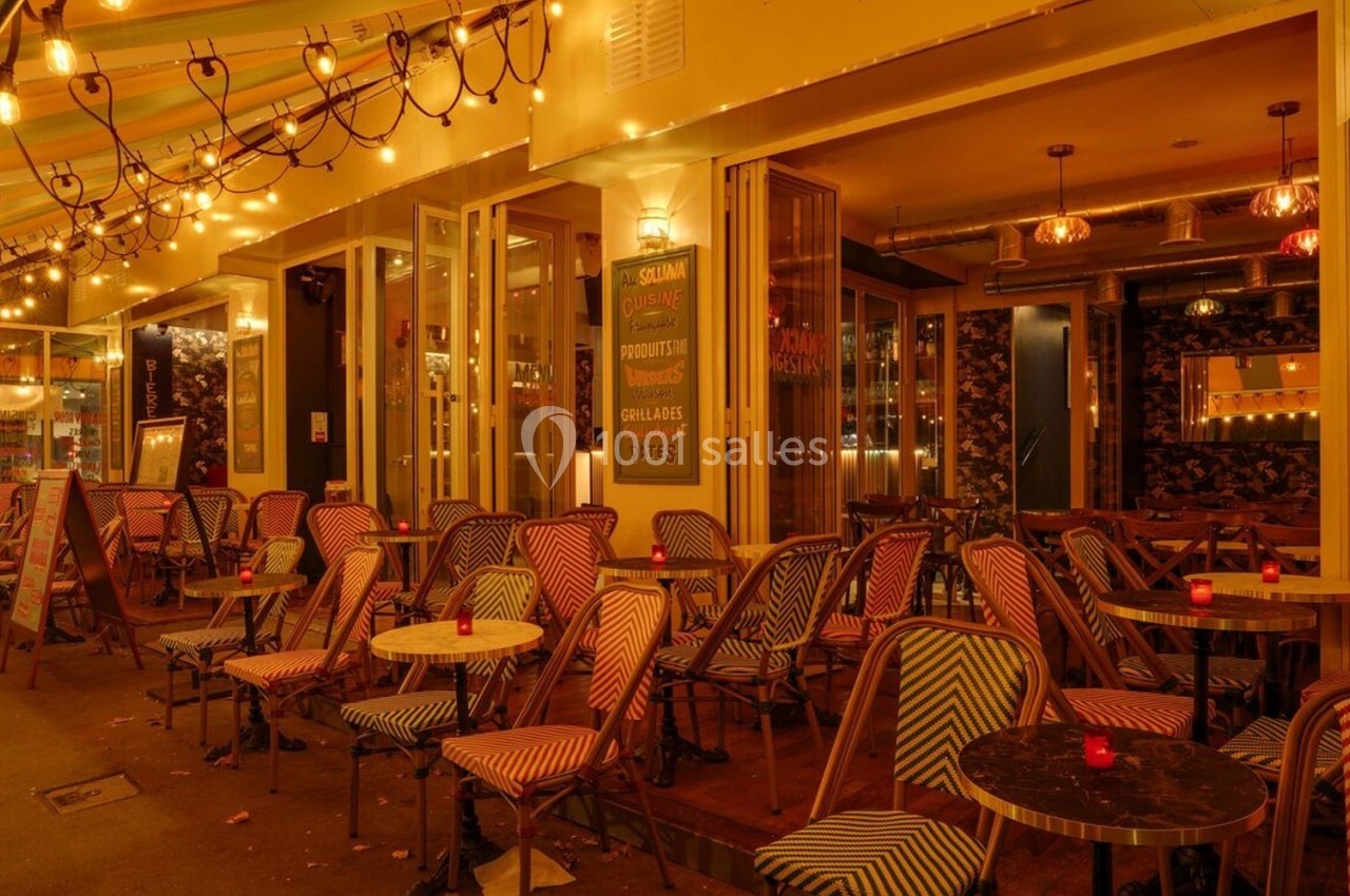 Terrasse d'un café-restaurant éclairée par des guirlandes lumineuses, avec des chaises colorées et des tables en soirée.