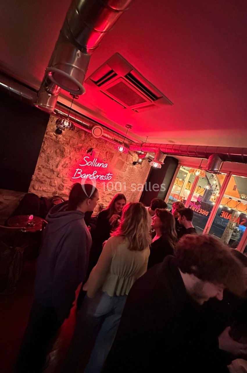 Groupe de personnes discutant dans un bar à l'éclairage rouge avec un mur en pierre et une enseigne lumineuse.