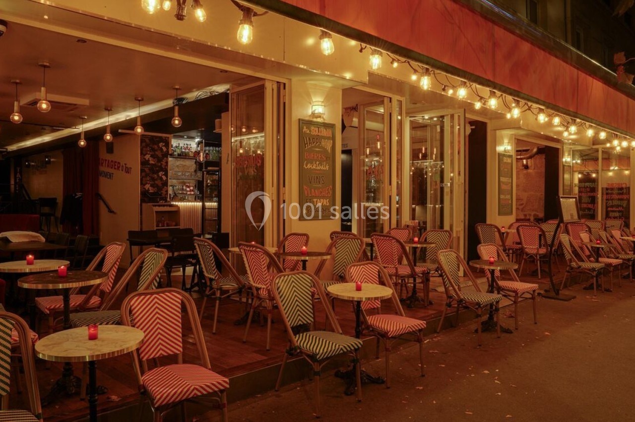 Terrasse d’un café-restaurant éclairée par des guirlandes lumineuses, avec des chaises et tables vides en soirée.