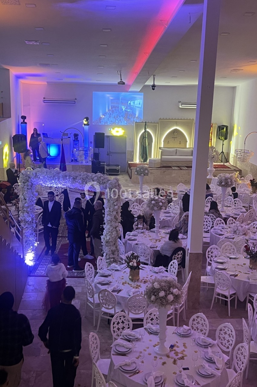 Salle de réception décorée pour un événement, avec tables dressées, arche fleurie et éclairage coloré.