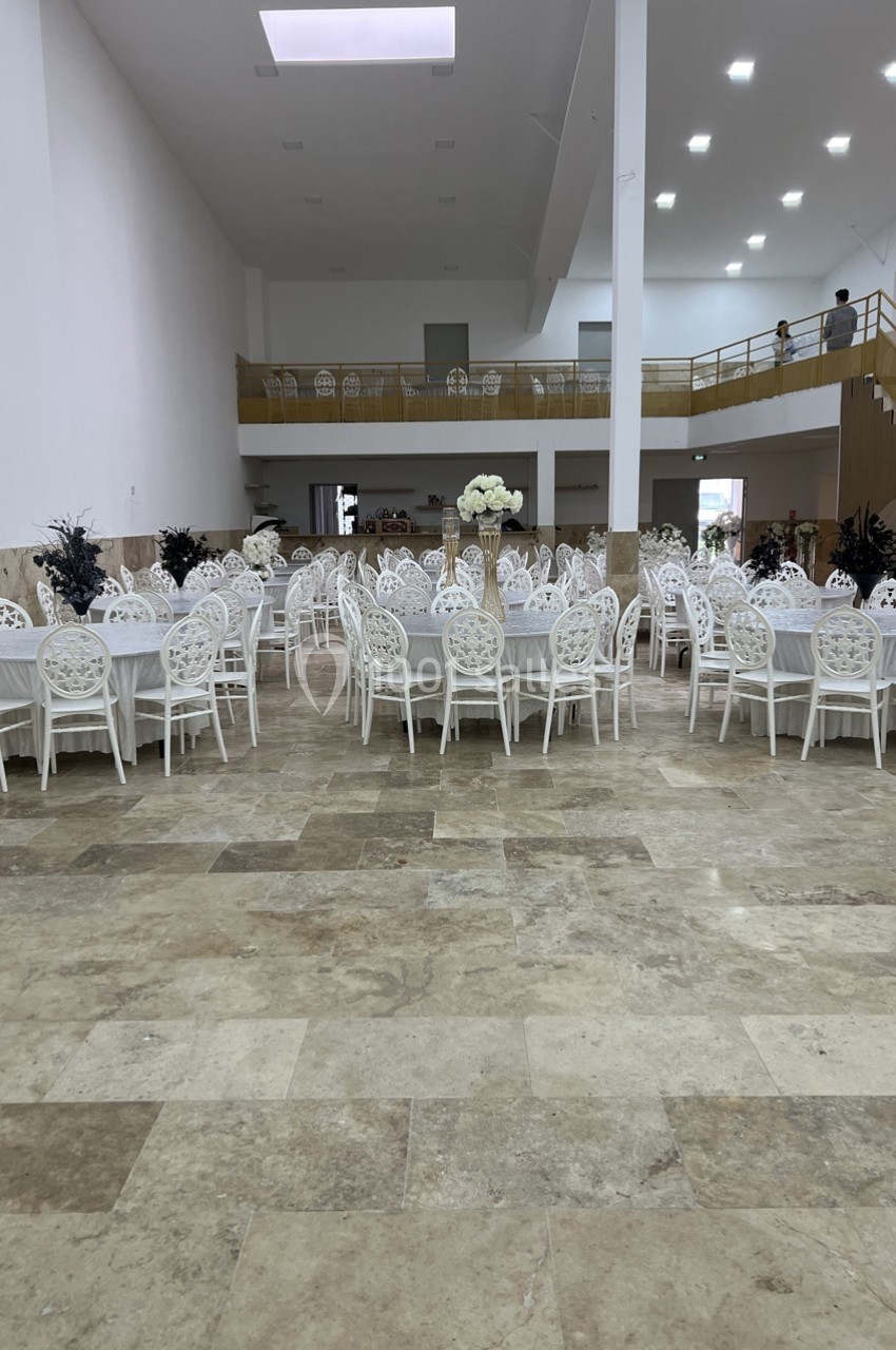 Salle spacieuse avec tables et chaises blanches, sol en pierre beige et mezzanine en arrière-plan.