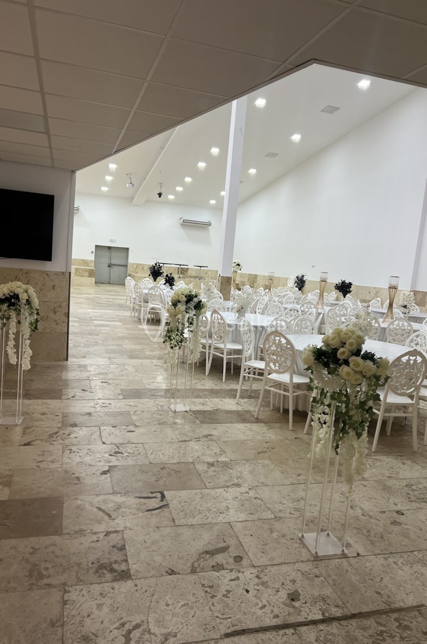 Salle de réception lumineuse avec tables blanches dressées et décorations florales élégantes sur sol en pierre.
