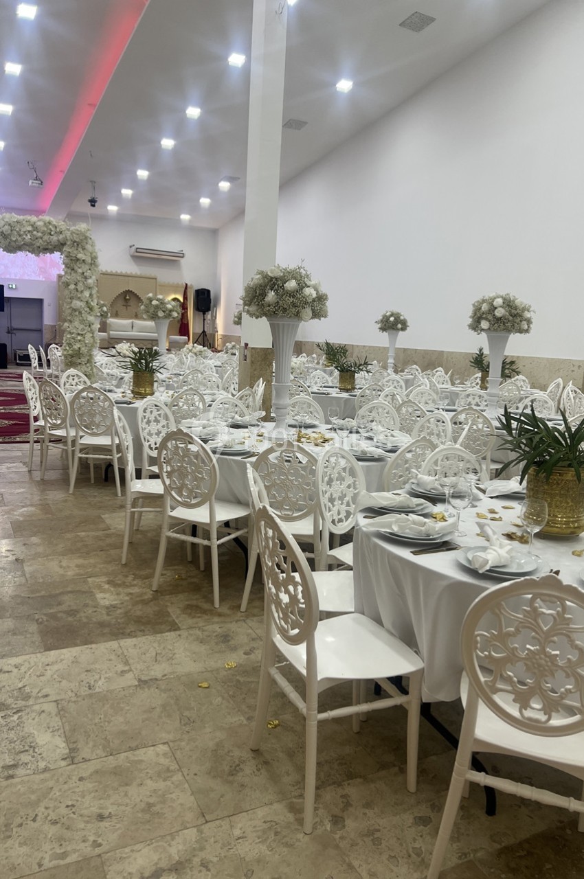 Salle de réception décorée avec des tables rondes, nappes blanches, chaises élégantes et arrangements floraux.