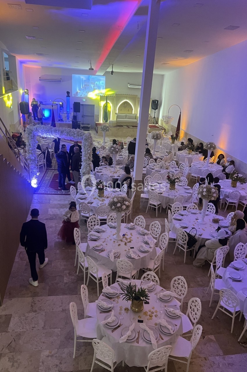 Salle de réception décorée pour un événement, avec des tables dressées et des invités assis ou debout.