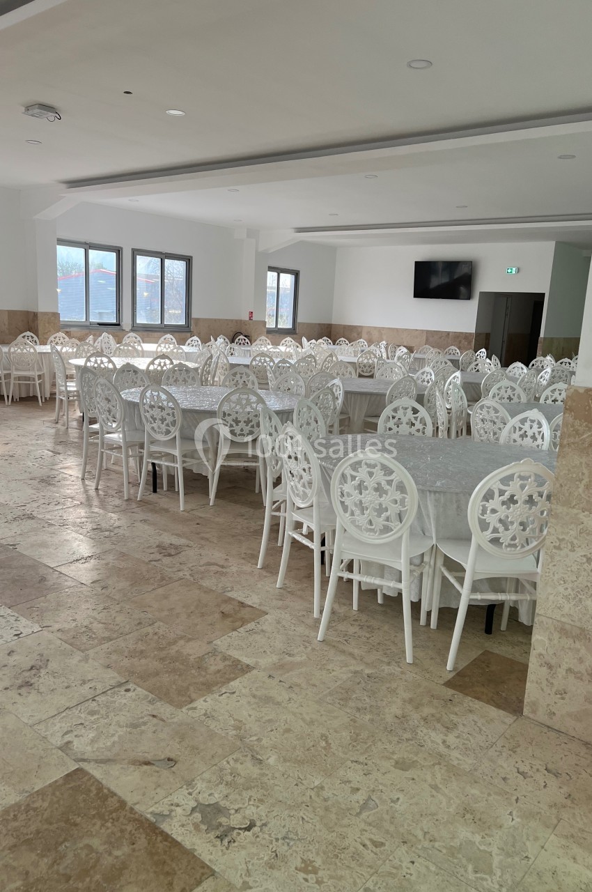 Salle spacieuse avec des tables rondes recouvertes de nappes blanches et des chaises blanches à motifs, éclairée par des…
