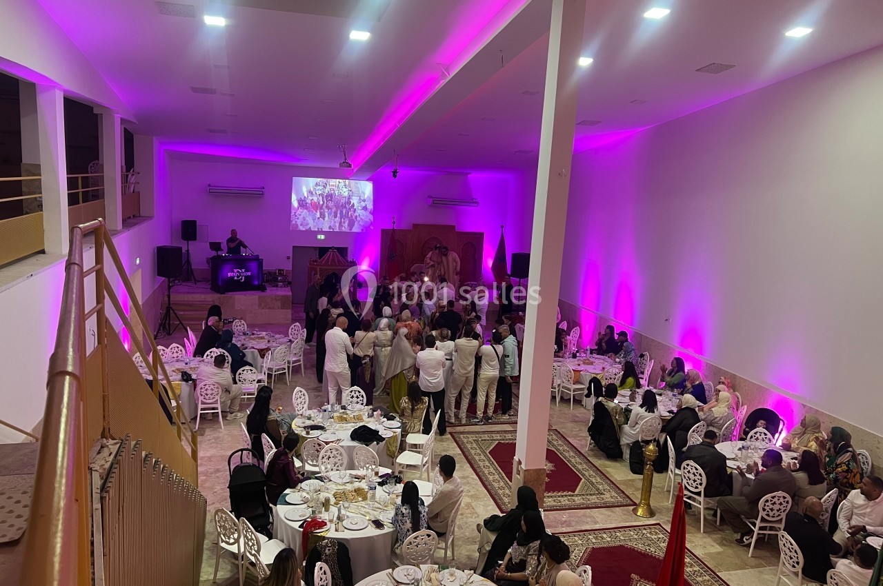 Salle de réception décorée de lumières roses, avec des invités assis et debout autour de tables lors d'un événement.