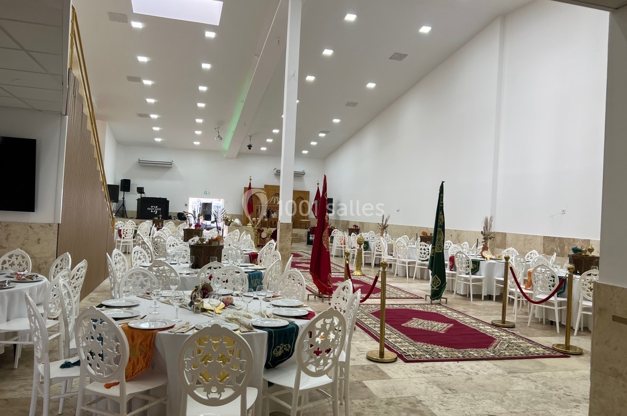 Salle de réception lumineuse avec tables dressées, chaises blanches décorées et tapis rouges dans un espace spacieux.