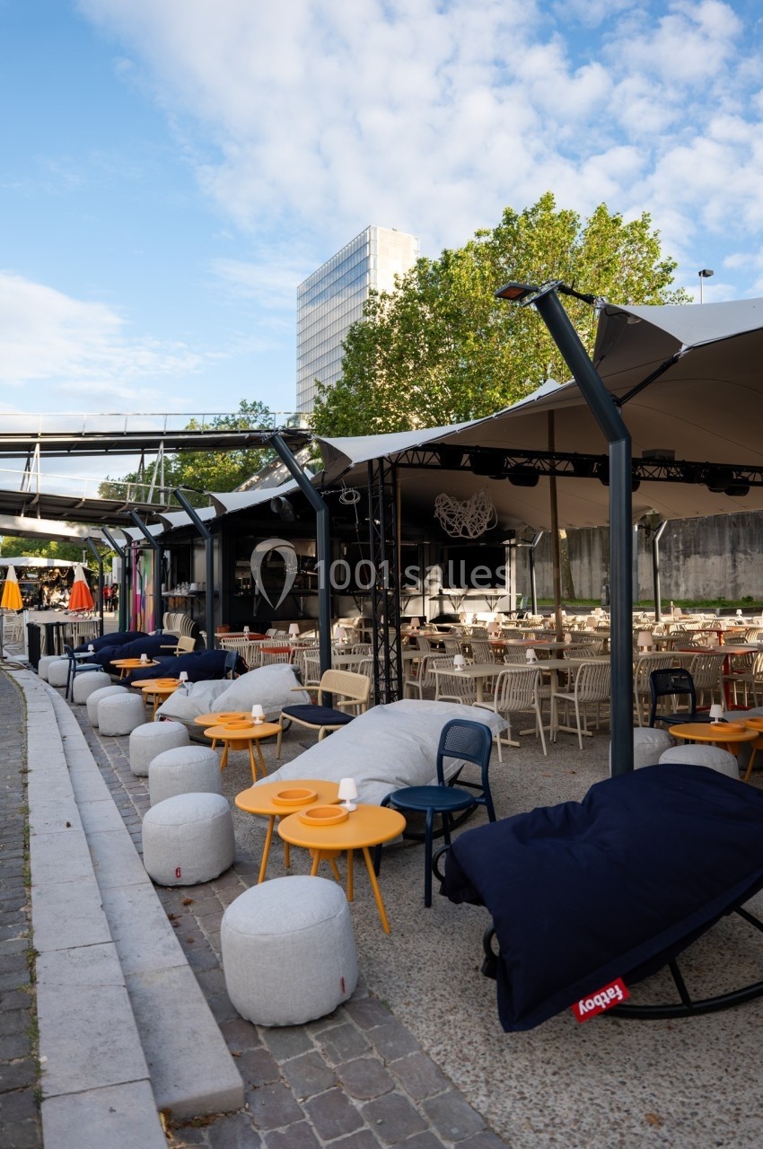 Terrasse extérieure avec tables, chaises et poufs colorés sous des parasols, entourée de verdure et bâtiments en arrière…