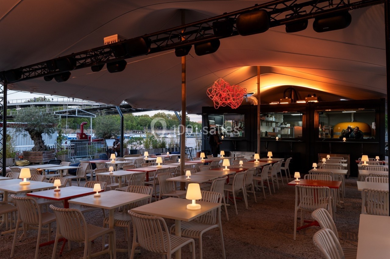 Terrasse de restaurant en soirée avec tables éclairées par des lampes, située près d'un cours d'eau.