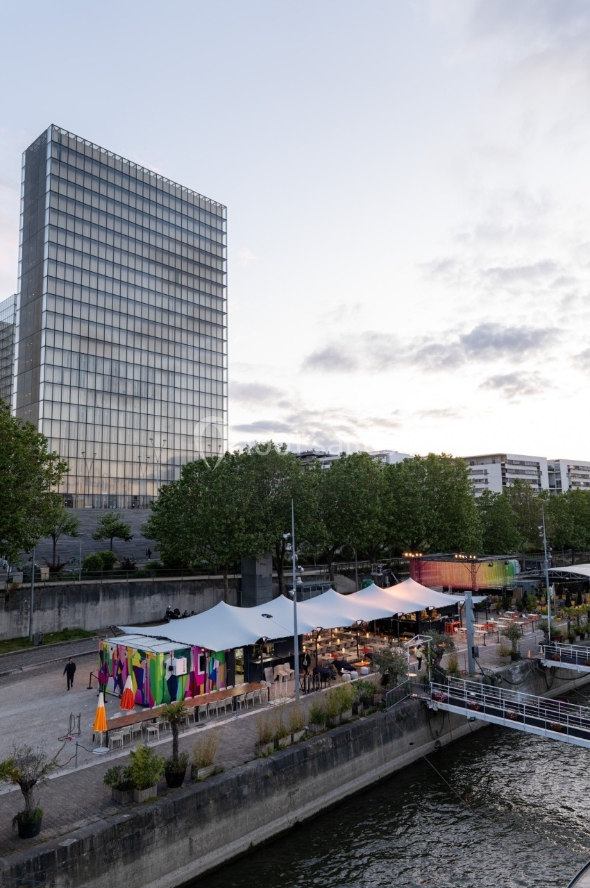 Vue d'un quai animé avec des stands colorés sous des tentes, près d'un bâtiment moderne et d'un cours d'eau.