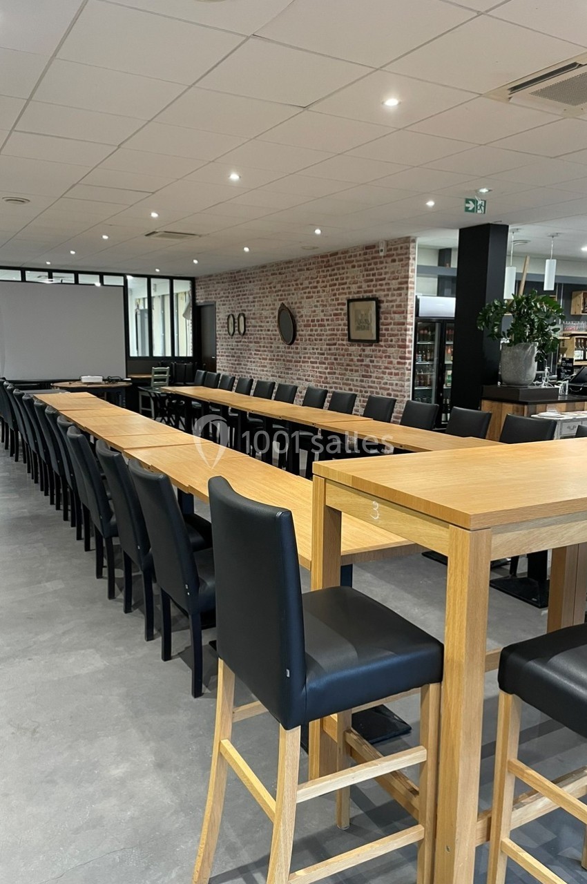 Salle de réunion avec longues tables en bois, chaises noires, écran de projection et décor moderne en briques.