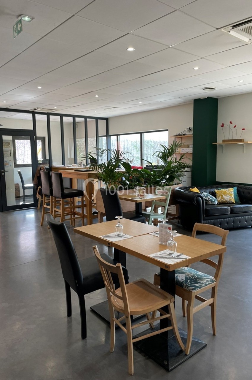 Salle lumineuse avec tables en bois, chaises variées, canapé noir, plantes et grande baie vitrée.