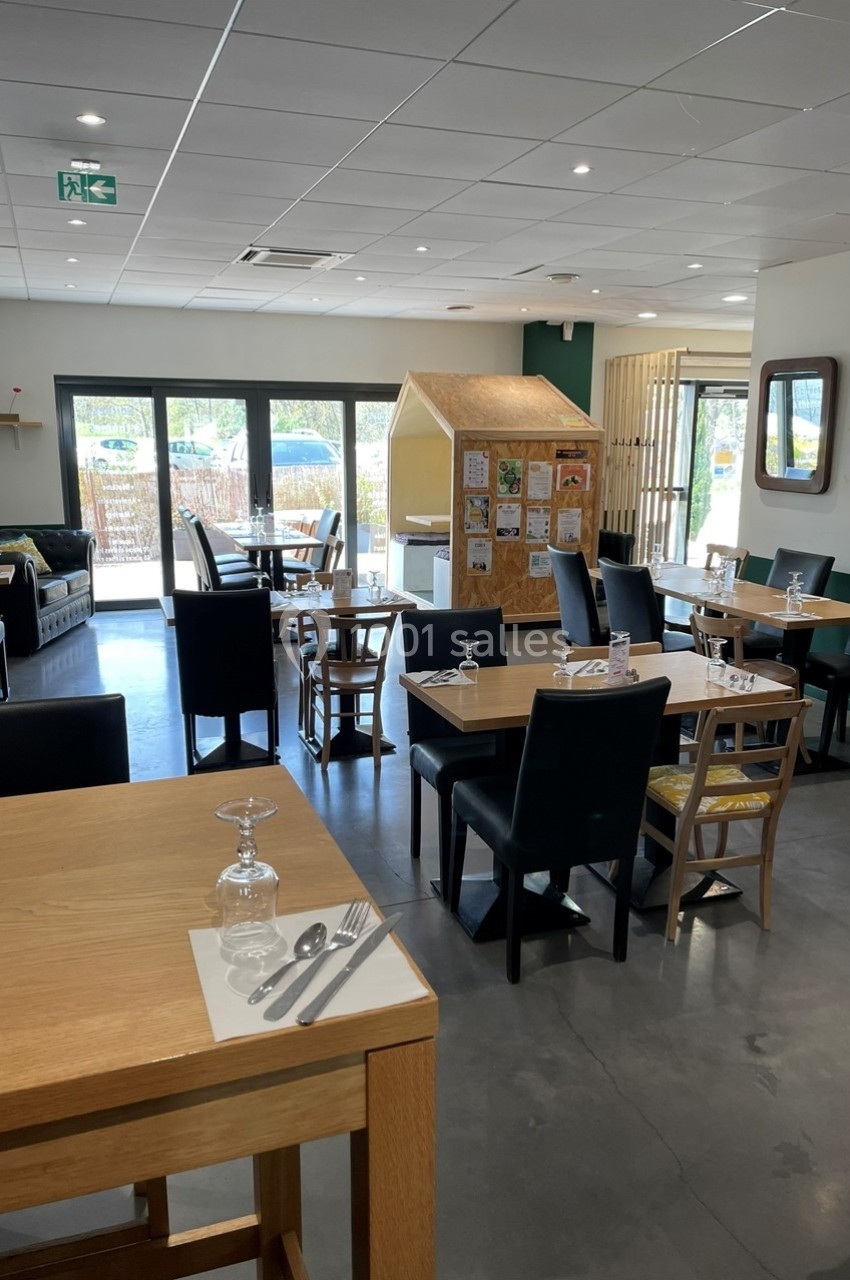 Salle de restaurant lumineuse avec tables en bois dressées, grandes baies vitrées et espace enfant en arrière-plan.