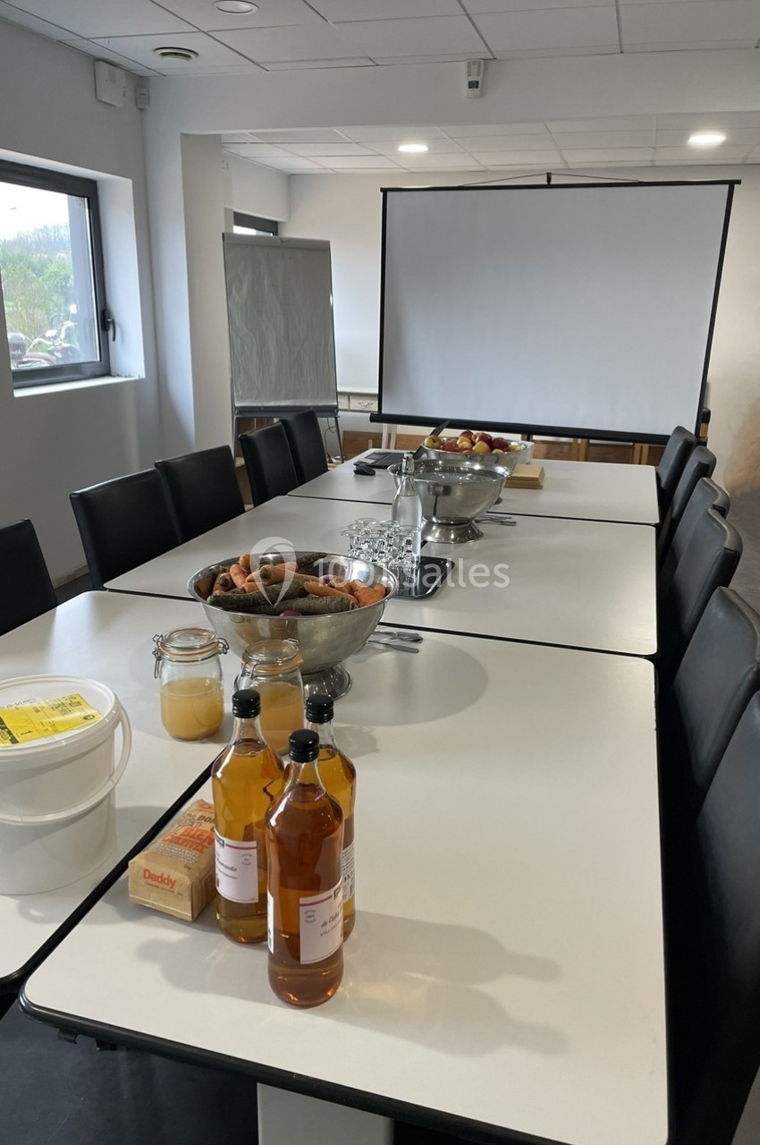 Salle de réunion avec une table dressée contenant des boissons, fruits, viennoiseries et matériel de présentation.