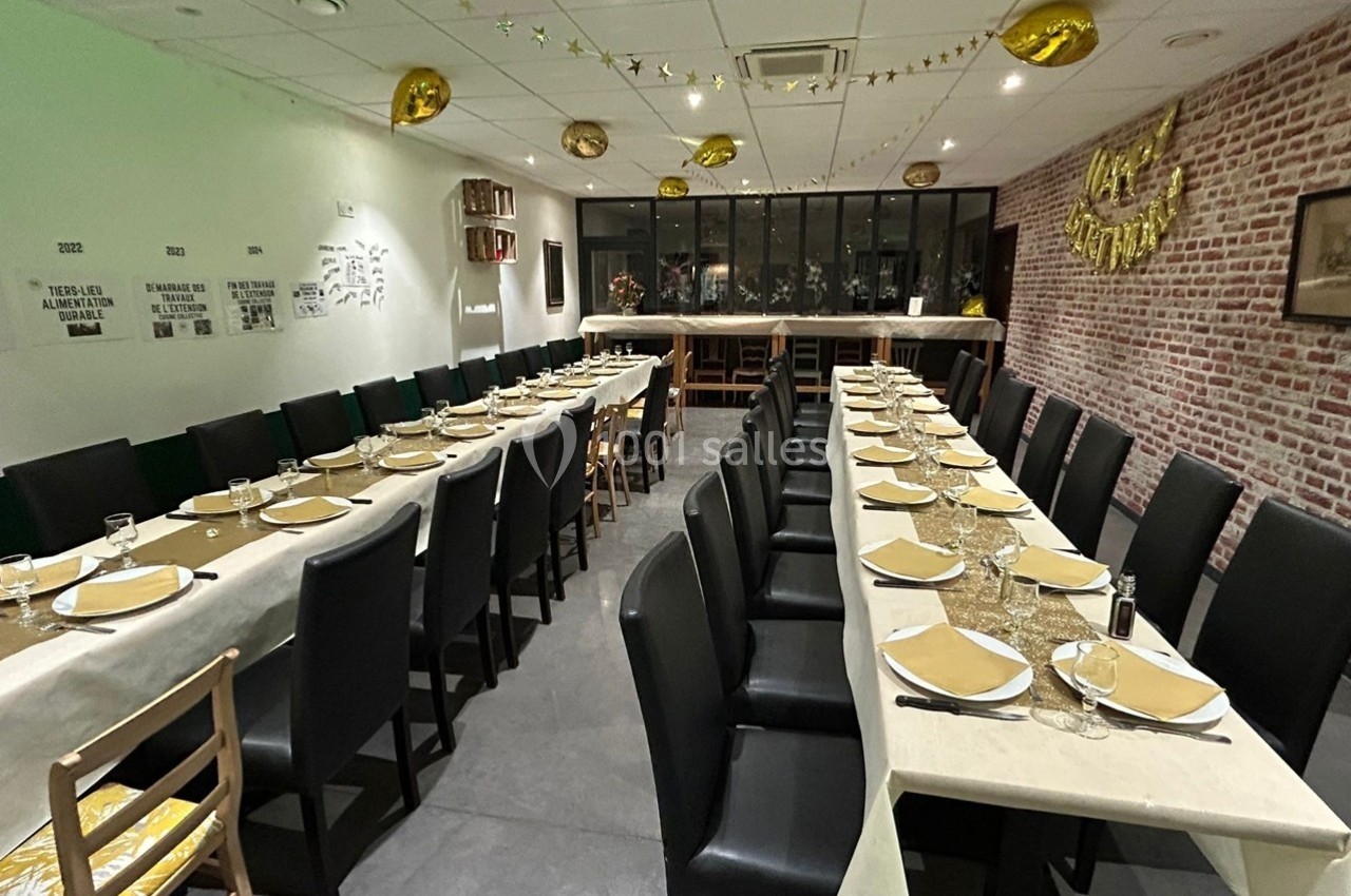 Salle décorée pour un événement, avec deux longues tables dressées, nappes blanches et ballons dorés suspendus.