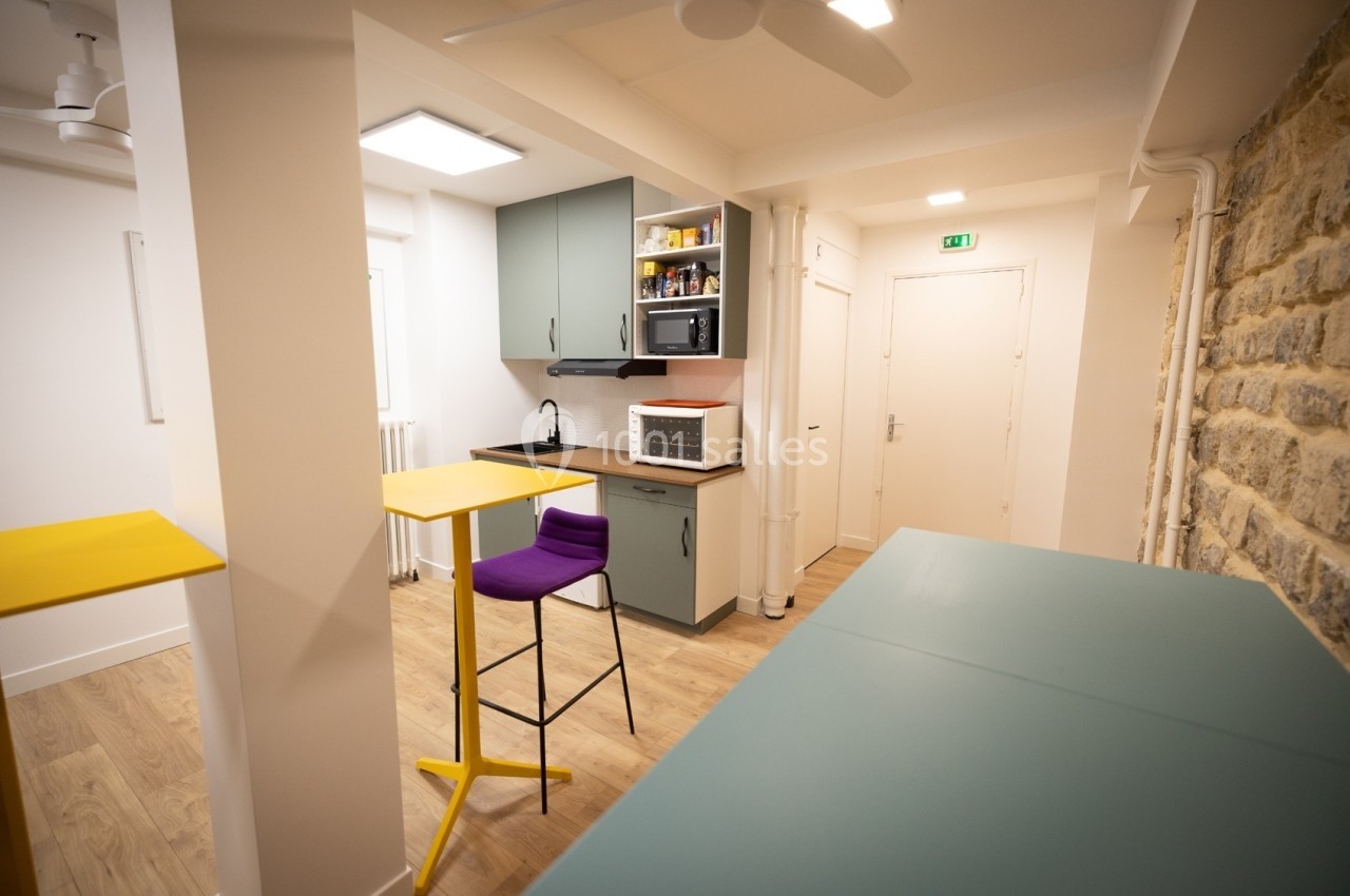 Cuisine moderne avec un mur en pierre, un plan de travail, des meubles verts et un tabouret violet près d'une table jaune.