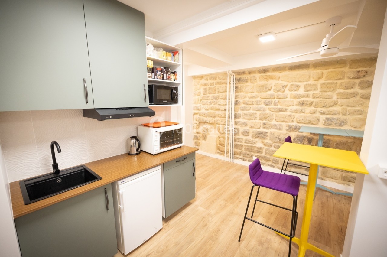 Cuisine compacte avec meubles verts, plan de travail en bois, mur en pierre apparente et table haute avec tabourets colorés.