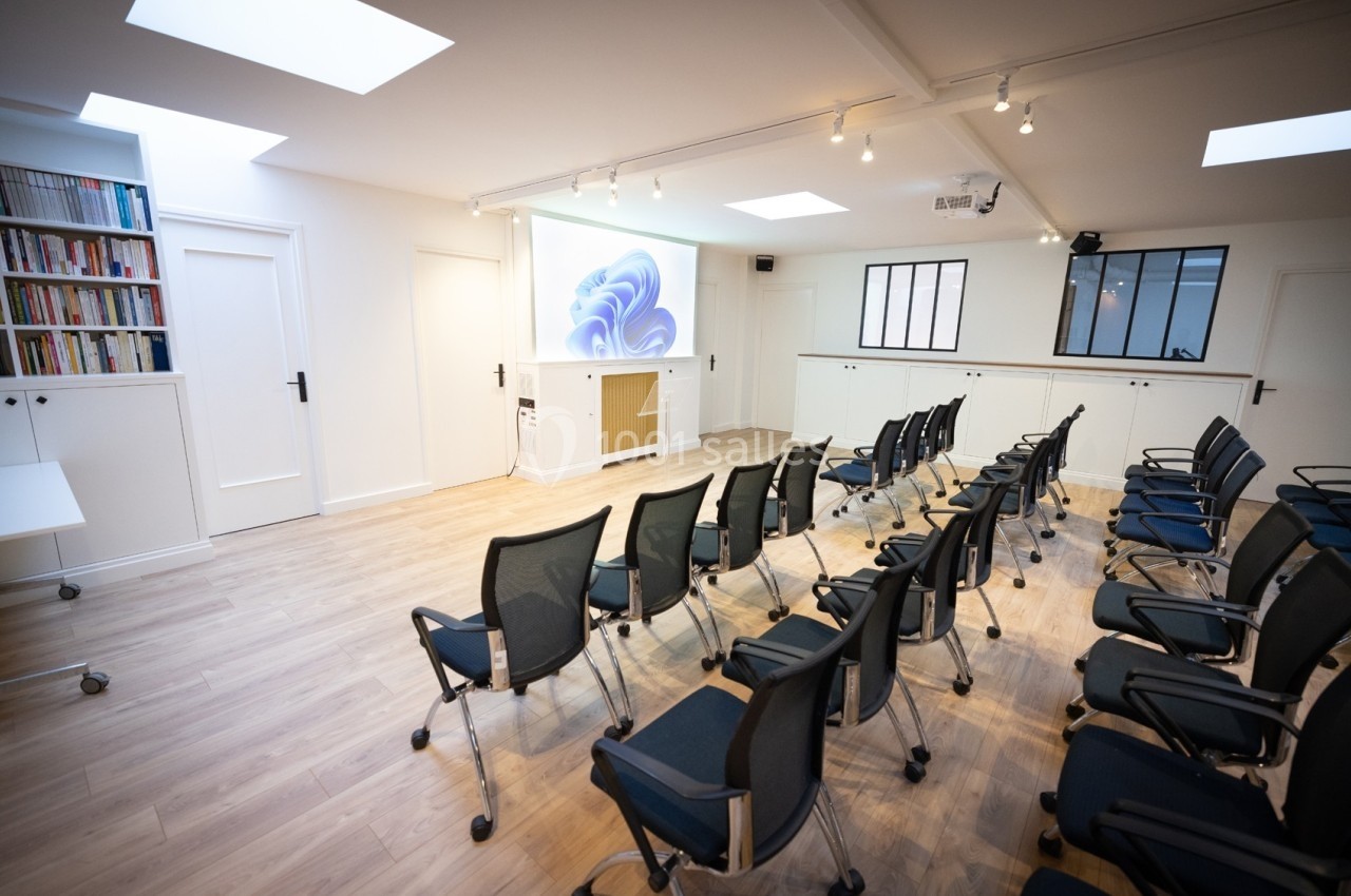 Salle lumineuse avec rangées de chaises noires, écran de projection allumé et étagères remplies de livres.