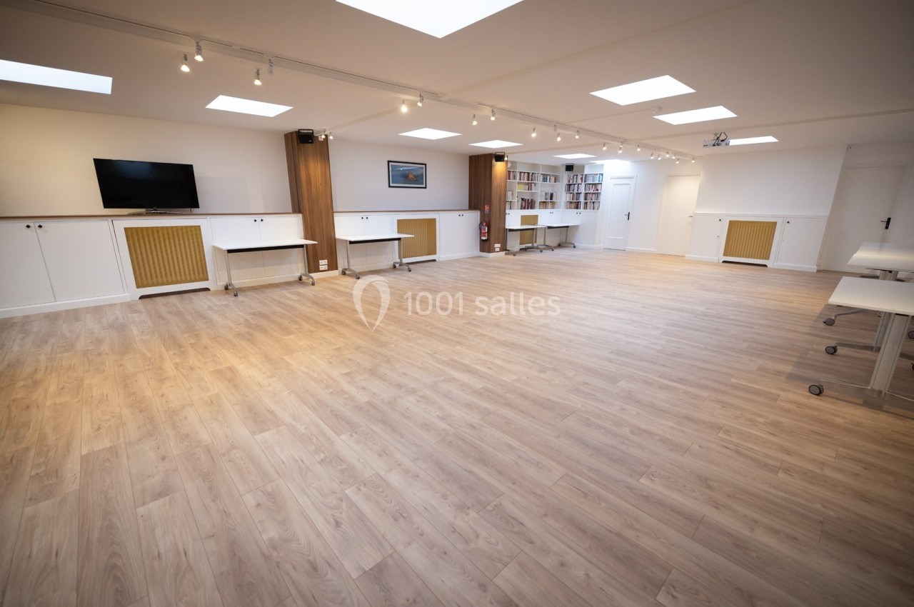 Salle lumineuse avec parquet clair, équipée de tables mobiles, étagères, écran mural et éclairage au plafond.