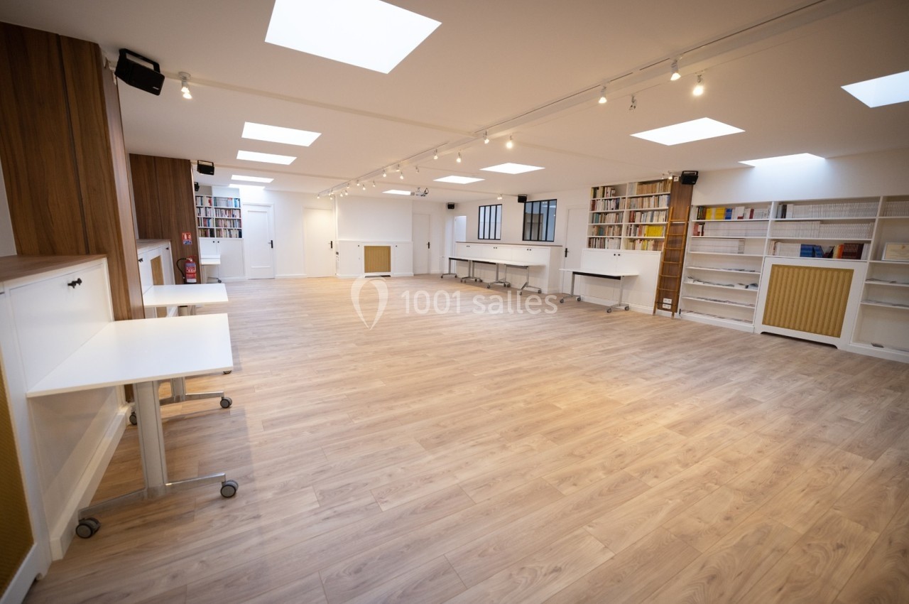 Salle lumineuse avec parquet clair, étagères remplies de livres, tables mobiles et éclairage au plafond.