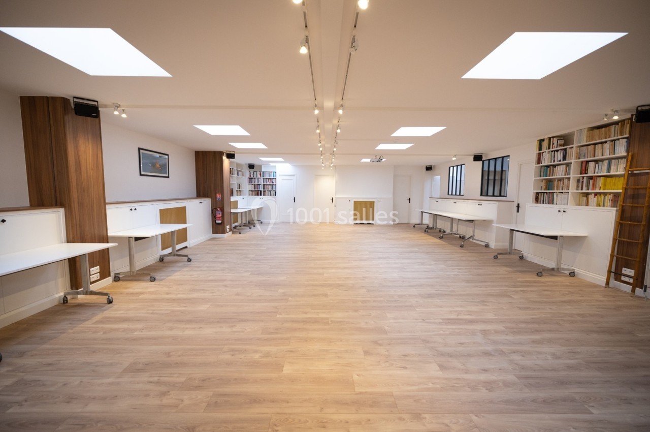 Salle lumineuse avec parquet clair, bureaux blancs sur roulettes, étagères remplies de livres et éclairage au plafond.