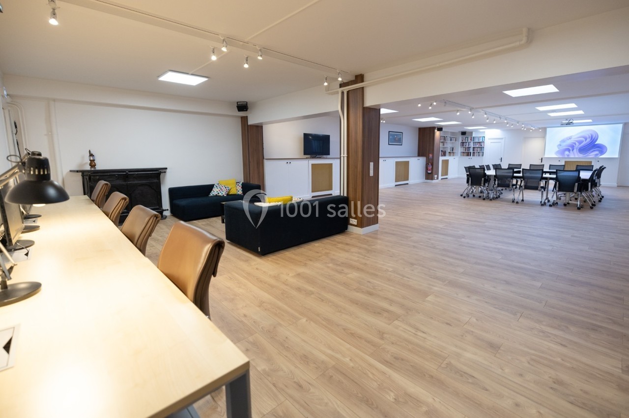 Salle spacieuse avec espaces de travail, canapé, table de réunion et bibliothèque, éclairée par des plafonniers.