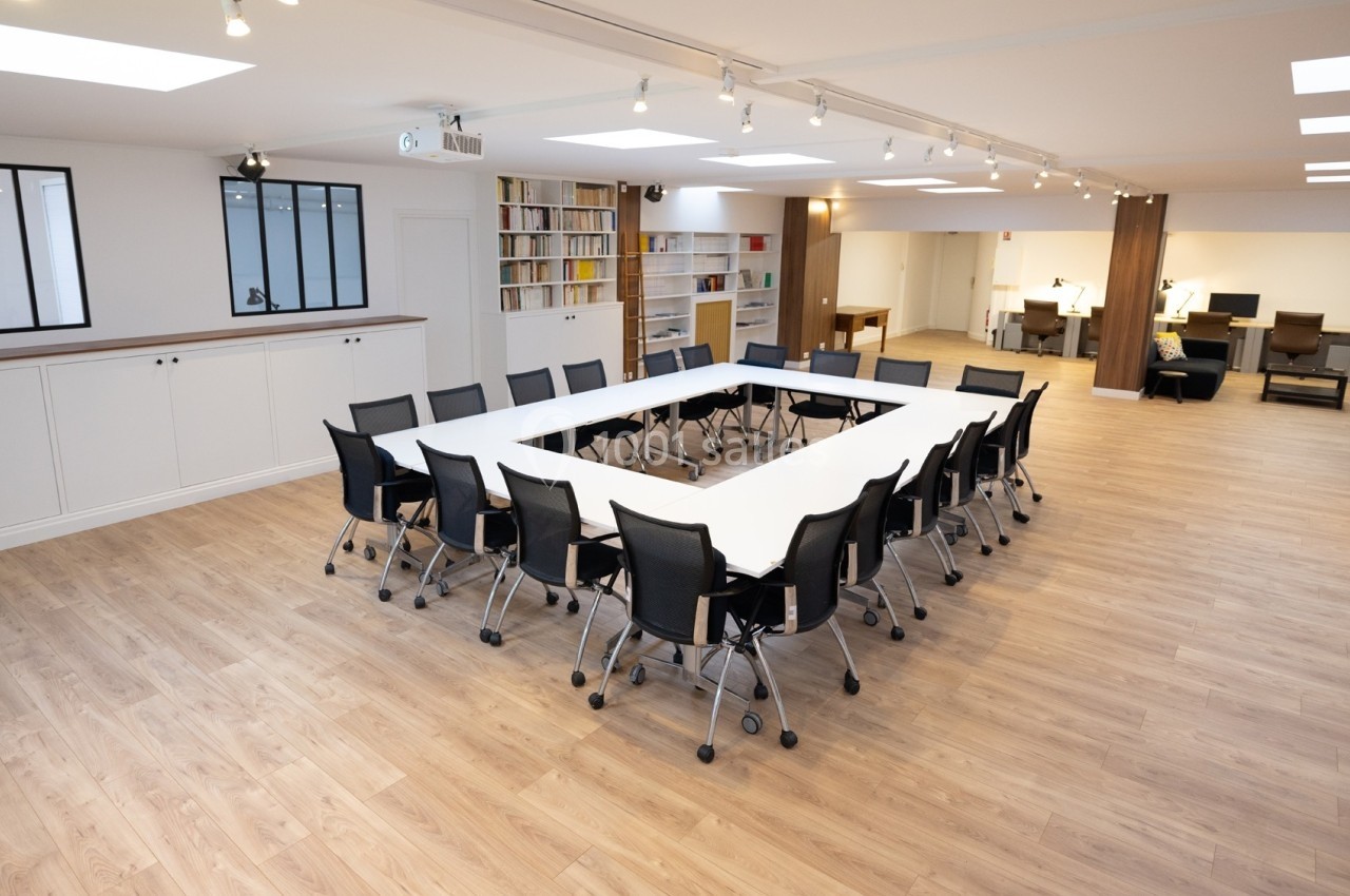 Salle de réunion lumineuse avec une grande table en U entourée de chaises noires, parquet clair et étagères en arrière-plan.