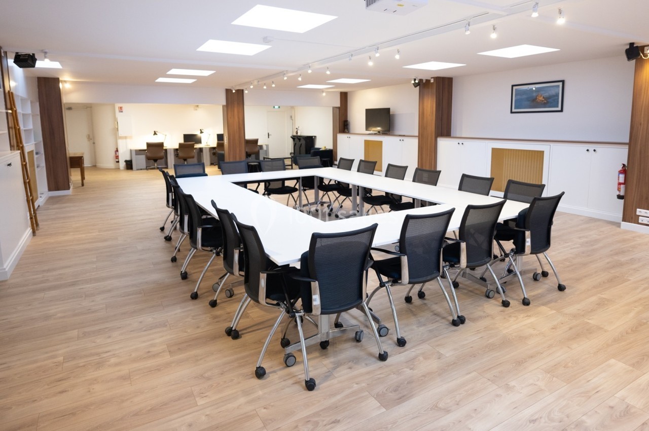 Salle de réunion lumineuse avec une grande table en U entourée de chaises noires sur un sol en bois clair.