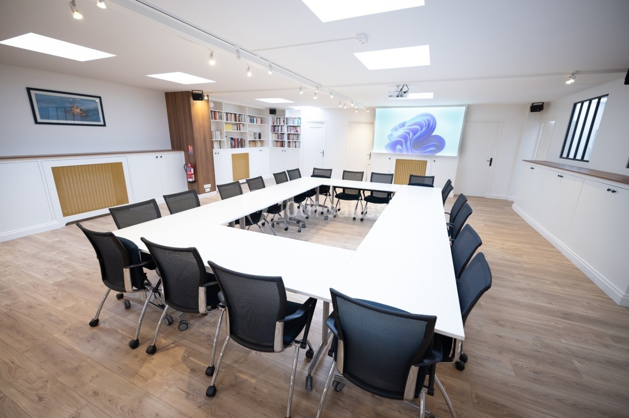 Salle de réunion lumineuse avec tables disposées en U, chaises noires, parquet clair et écran mural allumé.