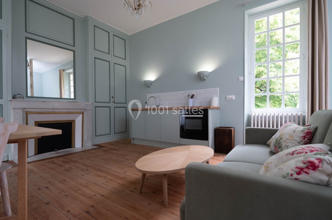 Salon lumineux avec parquet en bois, canapé, table basse, cheminée, cuisine ouverte et grande fenêtre donnant sur un jardin.
