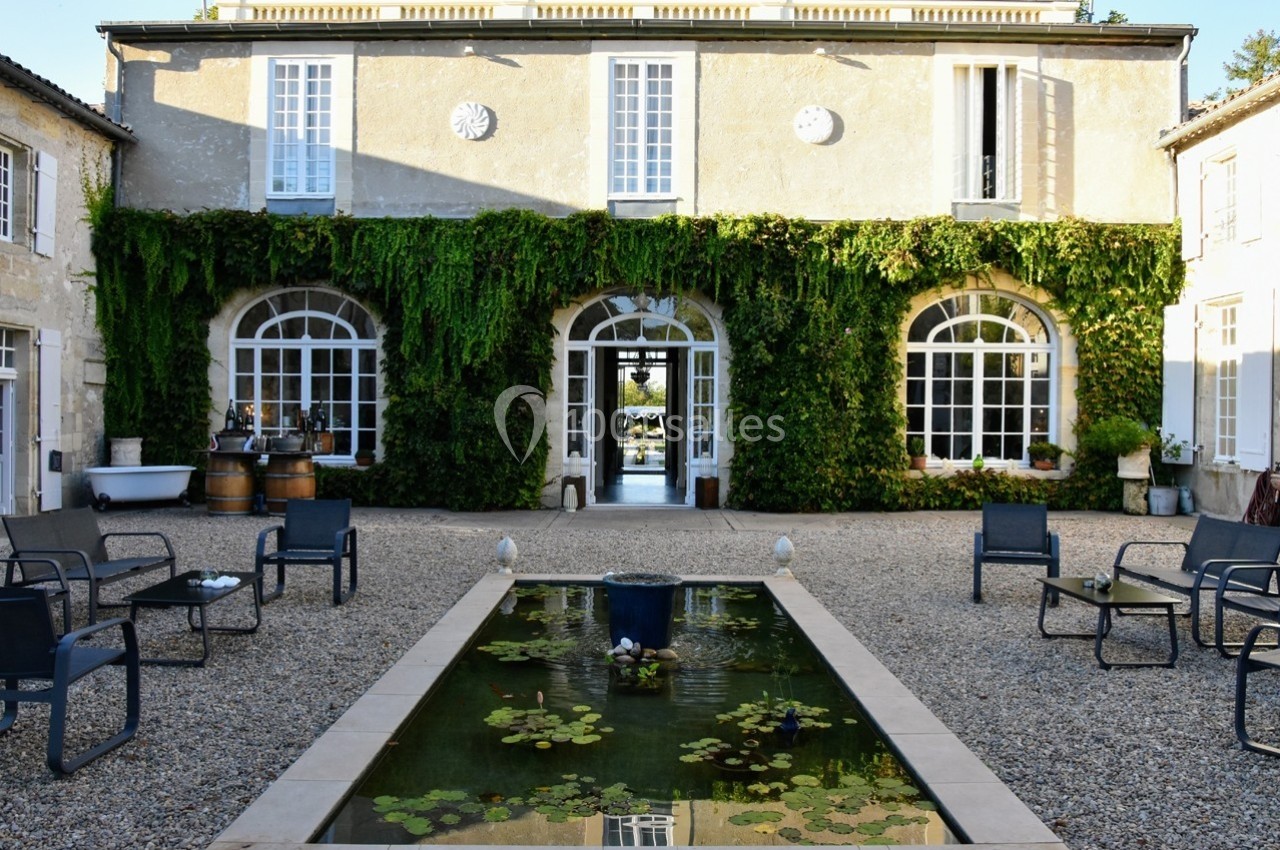Façade d'une maison en pierre recouverte de lierre, avec une cour, un bassin et des chaises disposées autour.
