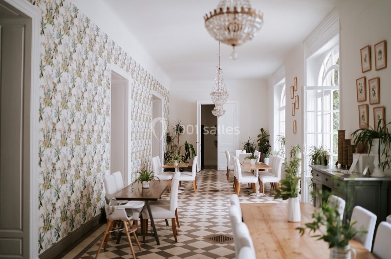 Salle lumineuse avec tables en bois, chaises blanches, plantes décoratives et lustres suspendus.