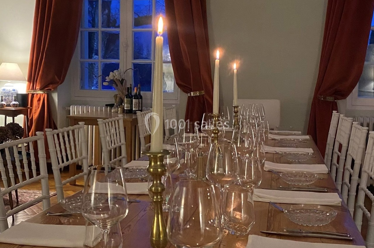 Table élégamment dressée avec chandeliers, verres et assiettes, dans une pièce éclairée par des bougies et des fenêtres.