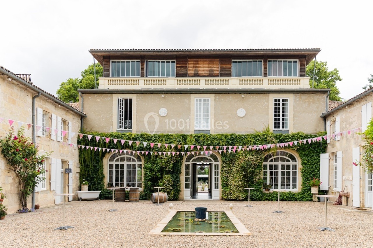 Façade d'une grande maison en pierre avec lierre, cour pavée, bassin central et guirlandes suspendues.
