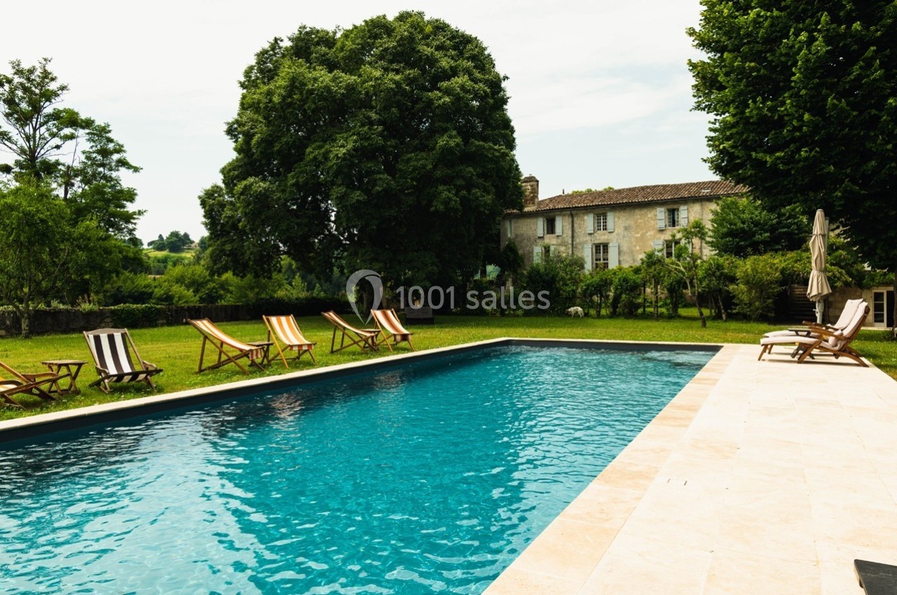 Piscine extérieure entourée de chaises longues et d'arbres, avec une maison en pierre en arrière-plan.