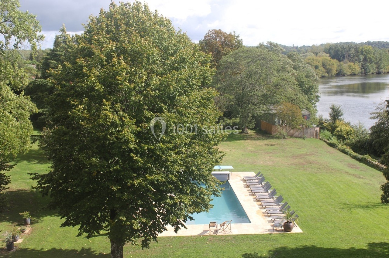 Vue d'un jardin avec piscine entourée de chaises longues, bordé par des arbres et une rivière en arrière-plan.