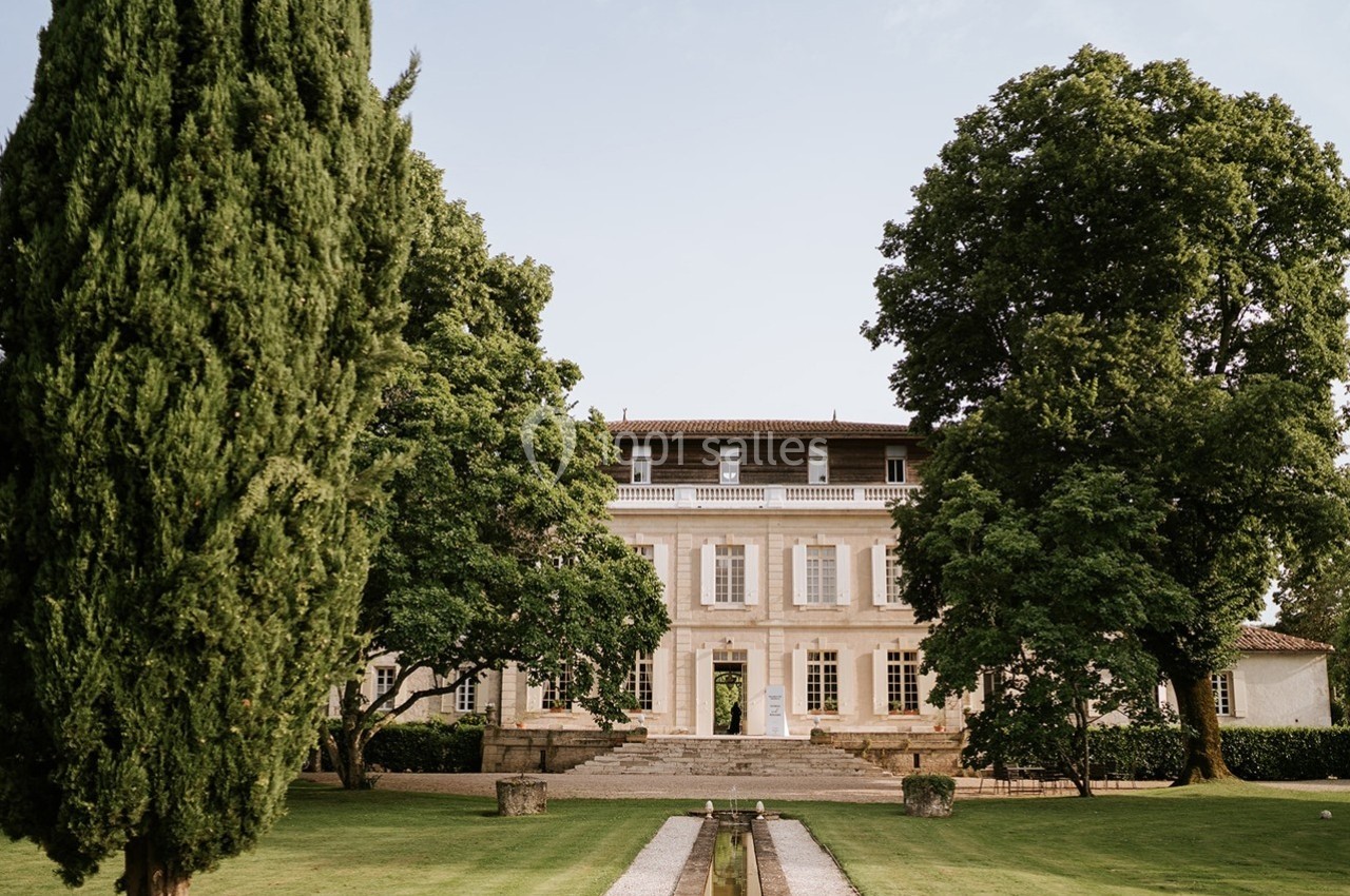 Façade d'un grand bâtiment ancien entouré de jardins verdoyants avec des arbres et une allée centrale.