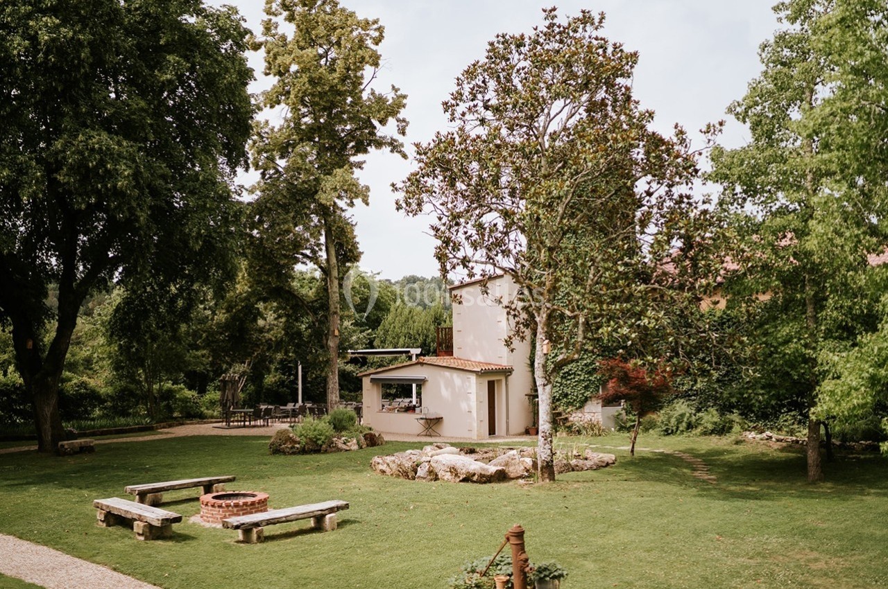 Jardin verdoyant avec pelouse, arbres, bancs en bois, foyer circulaire en pierre et petite maison en arrière-plan.